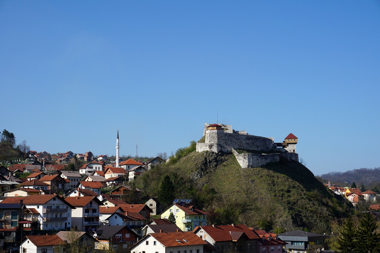 Kleinstadt mit roten Dächern; auf einem Hügel steht eine befestigte Burg, daneben ist ein schlankes Minarett unter klarem blauem Himmel.