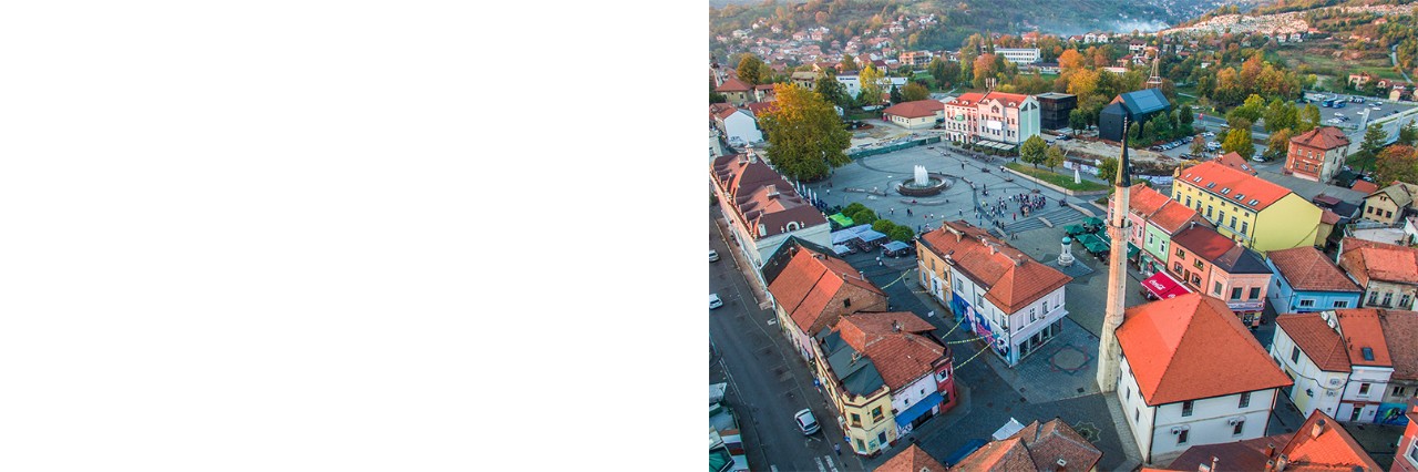 Blick von oben auf einen zentralen Platz mit Brunnen und Menschen, umgeben von bunten Häusern; im Vordergrund ist eine Moschee mit Minarett, im Hintergrund sind Hügel mit Herbstbäumen.