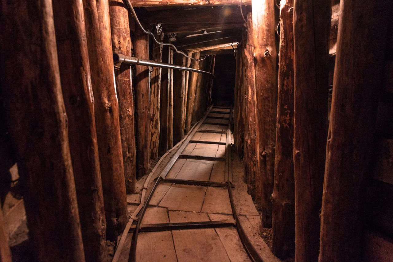 Unterirdischer Holztunnel mit seitlichen Stützbalken, sehr schmalem Laufweg, der ins Dunkle führt. Der Tunnel ist schwach beleuchtet und wirkt eng und niedrig.