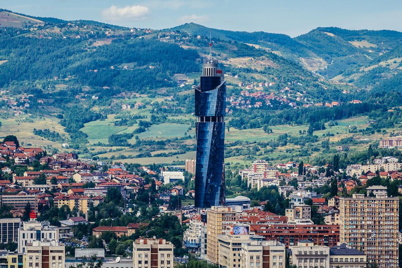 Hoher, moderner Glasturm mit verdrehter Form im Stadtgebiet von Sarajevo. Das Gebäude ragt über die umliegenden Häuser hinaus, im Hintergrund sind grüne Hügel und Wohnviertel zu sehen.