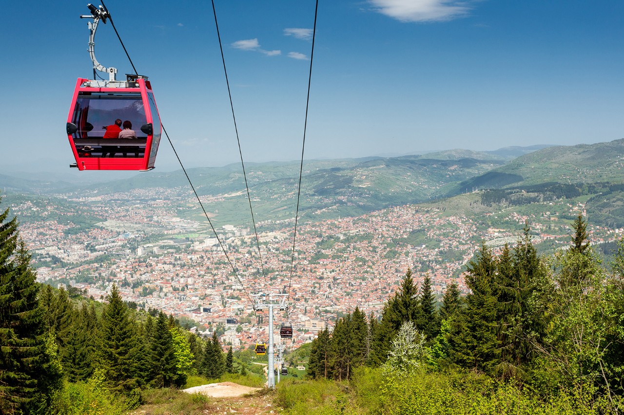 Rote Seilbahnkabine über bewaldetem Hang mit weiter Aussicht auf Sarajevo. Unterhalb erstreckt sich die Stadt in einem Tal, umgeben von grünen Bergen und blauem Himmel.