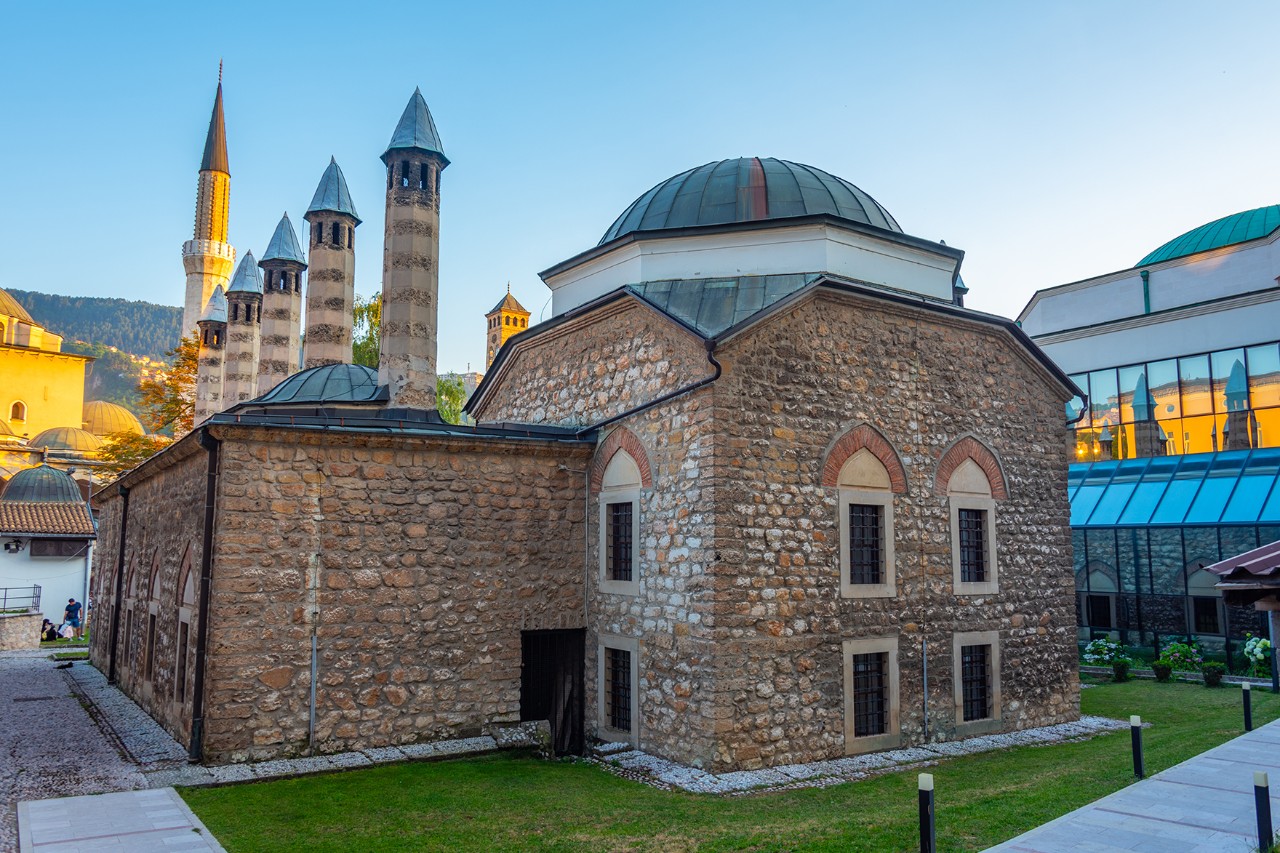 Historische Moschee aus Stein mit zentraler Kuppel und mehreren schlanken in Reihe stehenden Minaretten im Hintergrund. Das Gebäude befindet sich in einem Hofbereich mit Grünflächen und umliegenden Bauwerken.