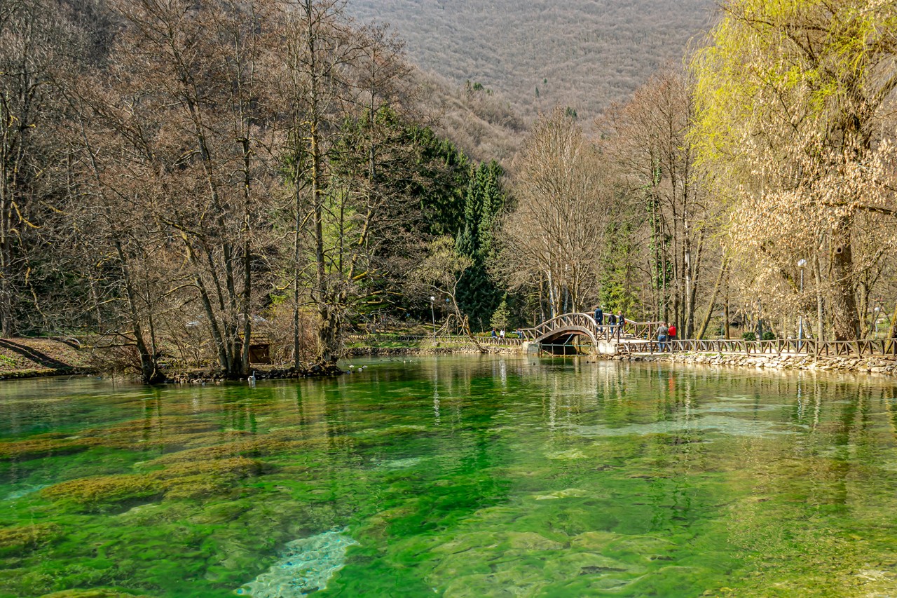 Klares, grün schimmerndes Quellwasser in einem naturbelassenen Parkgebiet mit noch kahlen Bäumen, Spazierwegen und einer kleinen Brücke, über die mehrere Menschen gehen. Im Hintergrund erheben sich bewaldete Hügel.