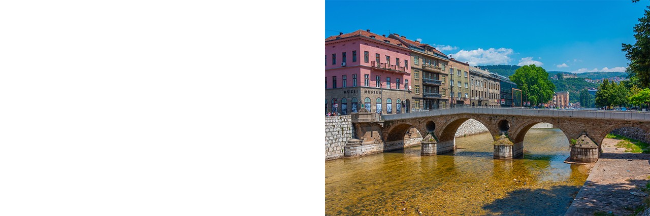 Eine historische Steinbogenbrücke überspannt einen Fluss. Am linken Ufer stehen dicht aneinandergereihte Häuser, im Hintergrund sind bewaldete Berge zu sehen.