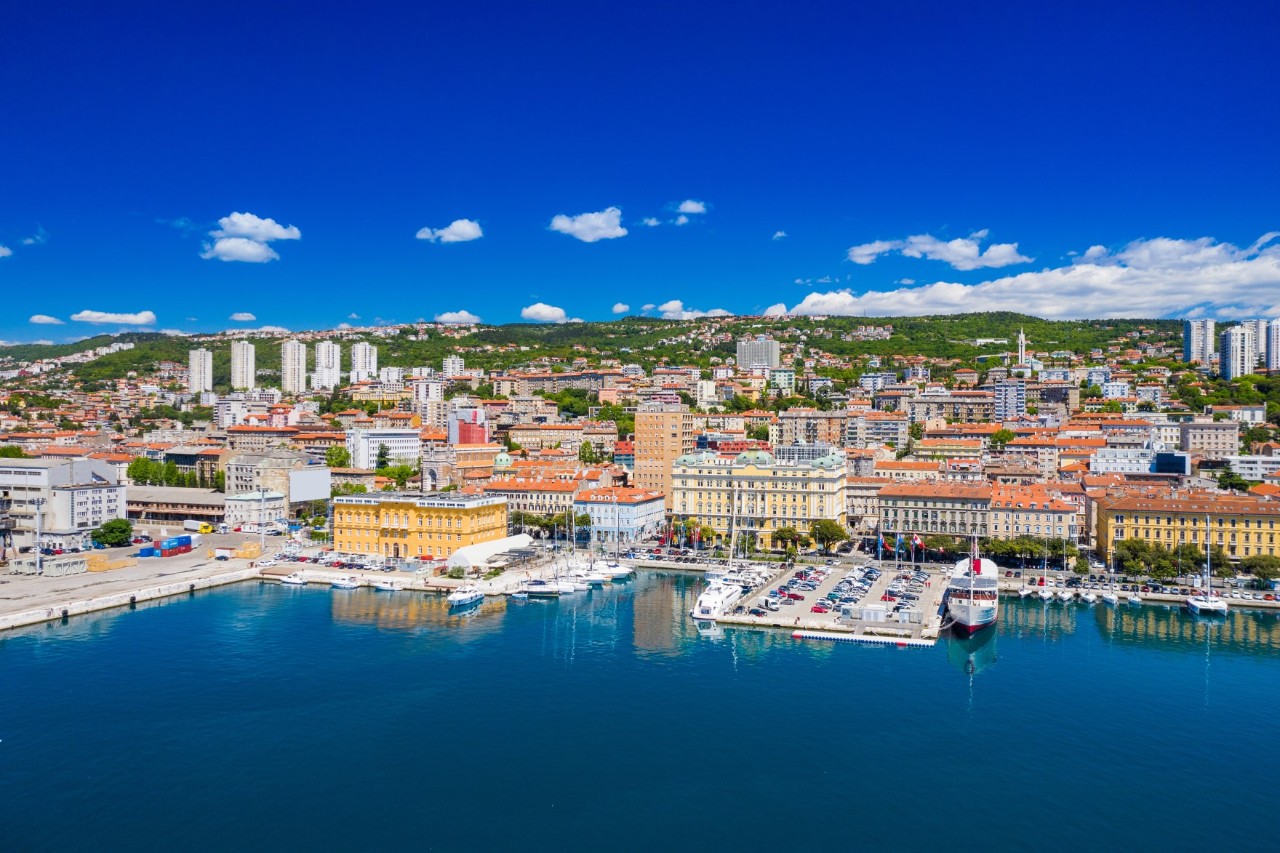 Altstadt von Rijeka mit Hafen, Wasserfront und grünen Hügeln im Hintergrund.