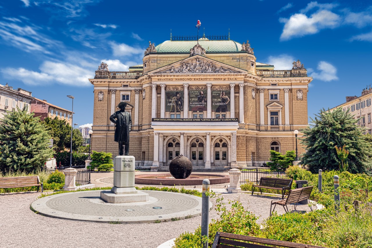 Kroatisches Nationaltheater in Rijeka mit Springbrunnen und Blumenbeeten davor.