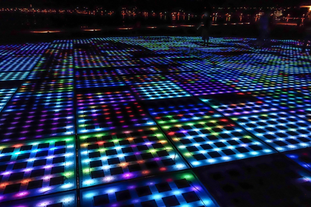 Ein farbiger Boden bei Nacht, auf dem verschiedenfarbige, einzelne Lichtelemente leuchten und die den Eindruck eines Mosaikmusters erwecken. Dahinter leuchten in der Ferne die Lichter der Stadt.