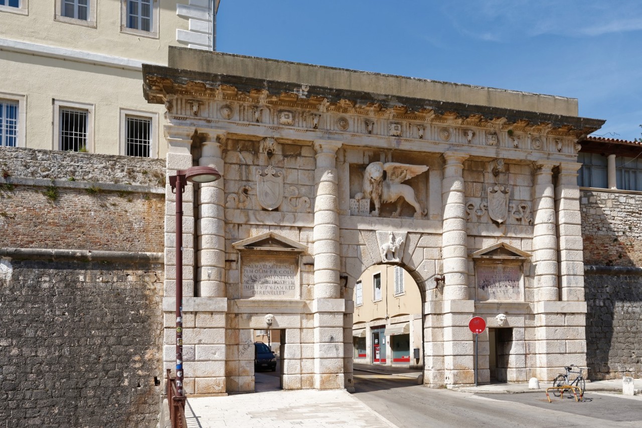Großes sandfarbenes, mit Säulen und Stuck verziertes Stadttor, durch das drei Tore führen. Das mittlere größte Tor ist bogenförmig, darüber ist ein großer, geflügelter Löwe angebracht. Über den anderen beiden Toren sind Wappen zu sehen. Links und rechts neben dem Tor ist eine Stadtmauer sichtbar, links erhebt sich ein großes, hellgelbes Gebäude.