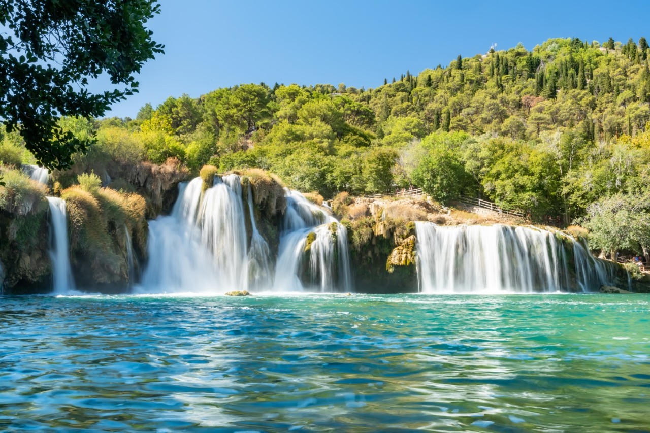 Mehrere Wasserfälle, die als Kaskaden über grüne, bewaldete Felsen in türkisfarbenes Wasser stürzen, umgeben von üppiger Natur.