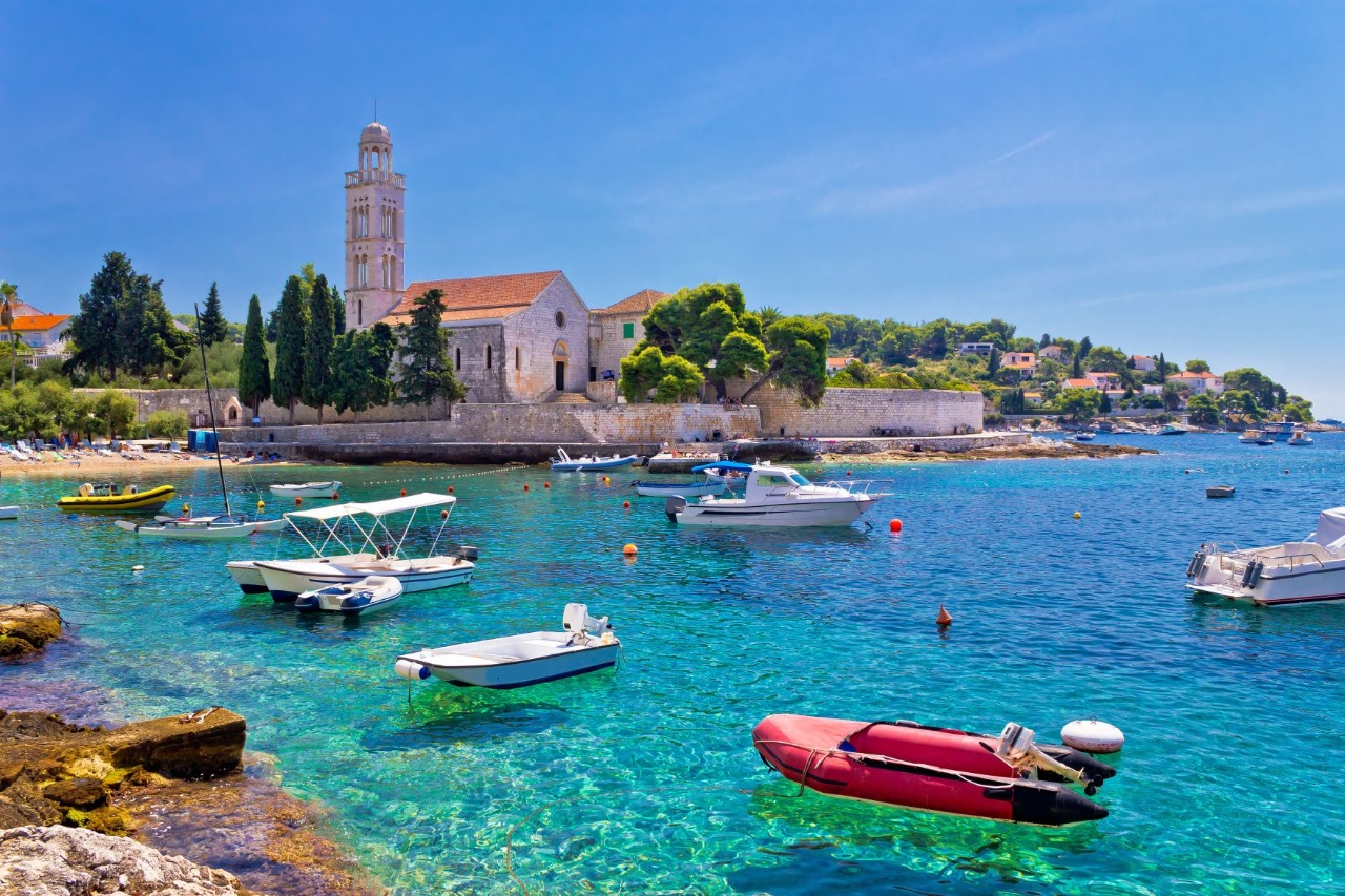 Küstenlandschaft mit kristallklarem Wasser, kleinen Booten und einer alten Kirche mit Glockenturm und mediterranen Bäumen im Hintergrund.