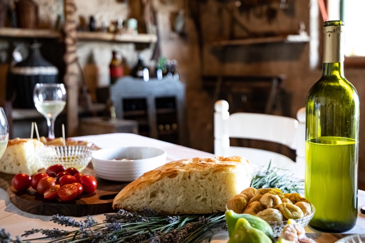 Rustikale Tischszene mit mediterranen Spezialitäten: frisches Brot, Kirschtomaten, getrocknete Feigen, ein Glas und eine Flasche Weißwein. Im Hintergrund ist eine Wand mit gefüllten Regalen zu sehen.