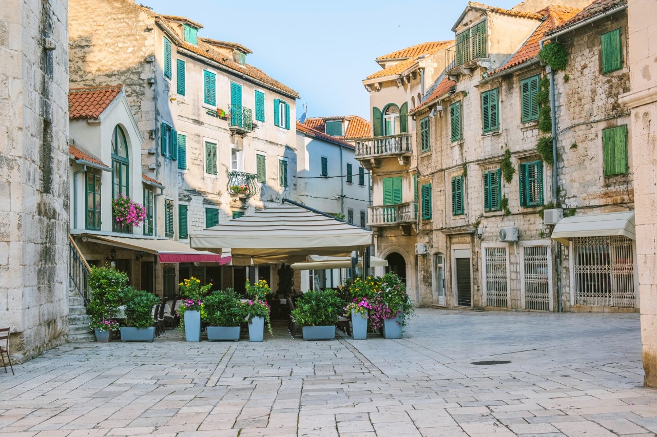 Platz in einer Altstadt mit alten Steinhäusern, grünen Fensterläden und roten Ziegeldächern. In der Mitte befindet sich ein Straßencafé mit Sonnenschirmen und Pflanzen. 