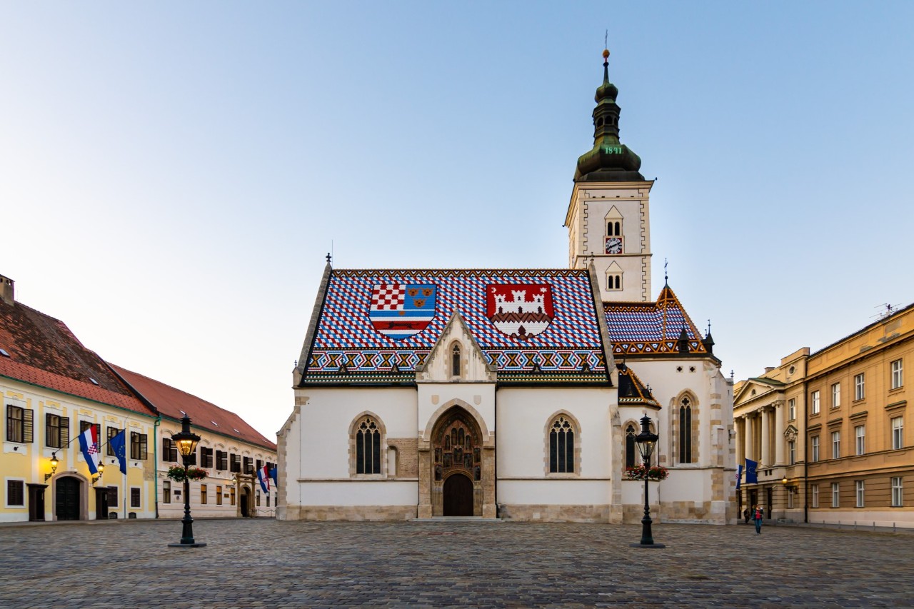 Kirche mit bunten Dachziegeln und zwei aufgemalten Wappen, umgeben von Häusern und menschenleerem Platz davor 