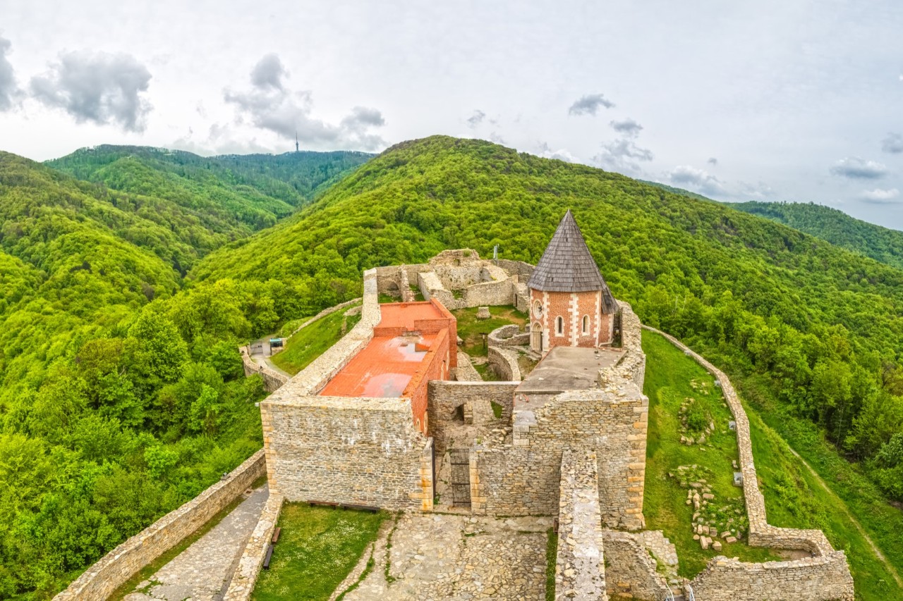Burg mit Mauern und Turm, umgeben von grüner Berglandschaft