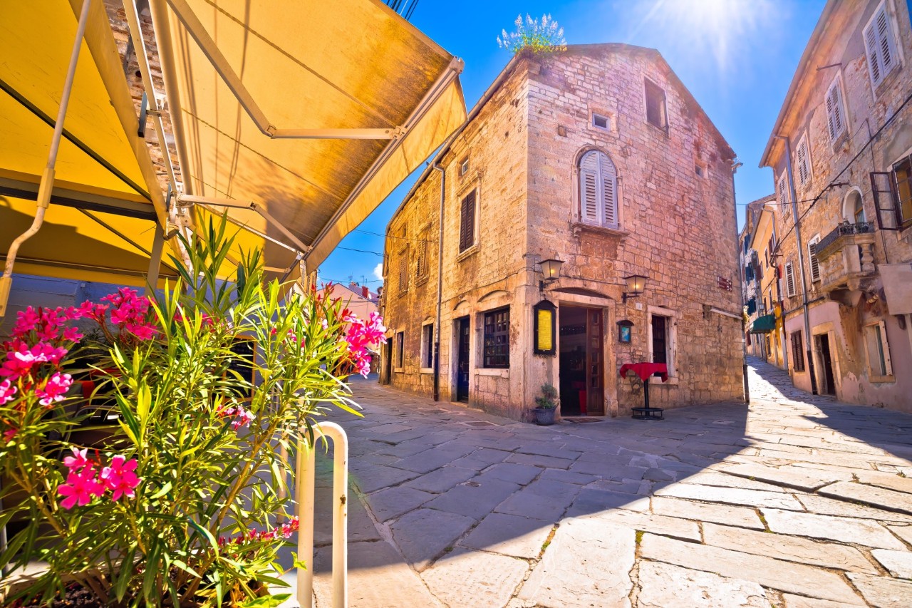 Eine gepflasterte Altstadtgasse mit historischen Steinhäusern im Sonnenschein. Im Vordergrund sind Oleanderblüten und Markisen zu sehen.