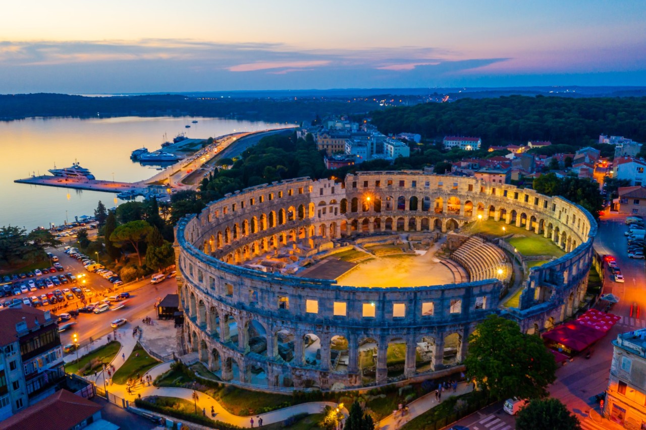 Ein beleuchtetes Amphitheater in der Abenddämmerung. Im Hintergrund sind die Küste und ein Hafen zu sehen, umgeben von Häusern und grüner Landschaft.