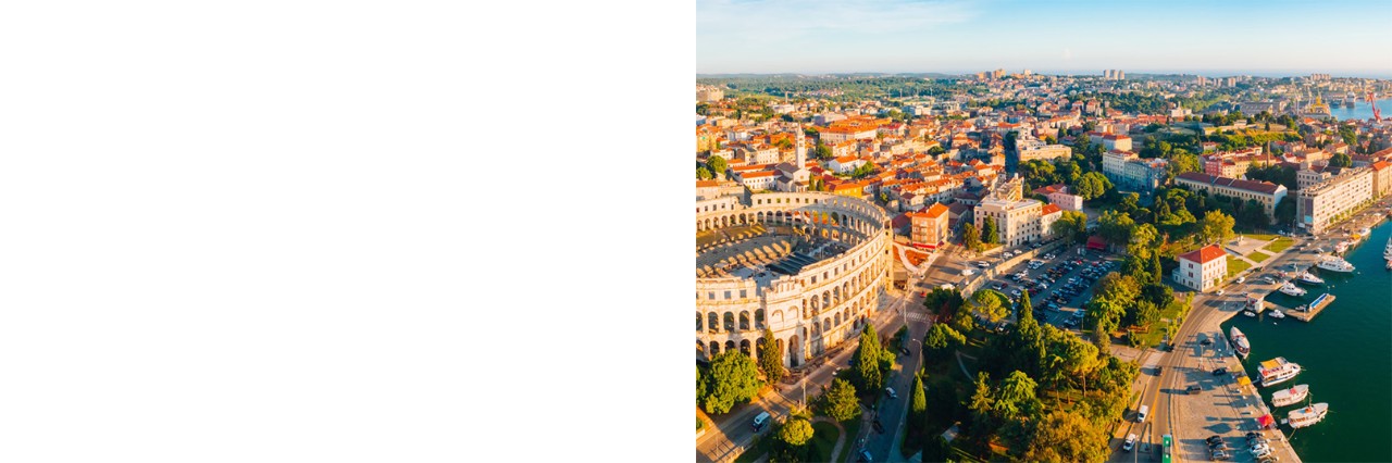 Luftaufnahme der Stadt Pula mit dem römischen Amphitheater links im Bild, während sich rechts eine Hafenpromenade mit Booten befindet. Im Hintergrund wird die Stadt mit ihren charakteristischen roten Dächern und grünen Parks sichtbar. 