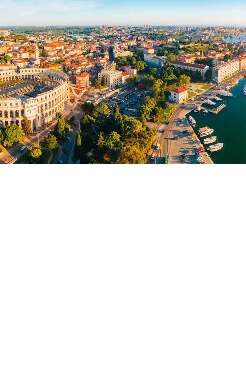 Luftaufnahme der Stadt Pula mit dem römischen Amphitheater links im Bild, während sich rechts eine Hafenpromenade mit Booten befindet. Im Hintergrund wird die Stadt mit ihren charakteristischen roten Dächern und grünen Parks sichtbar. 