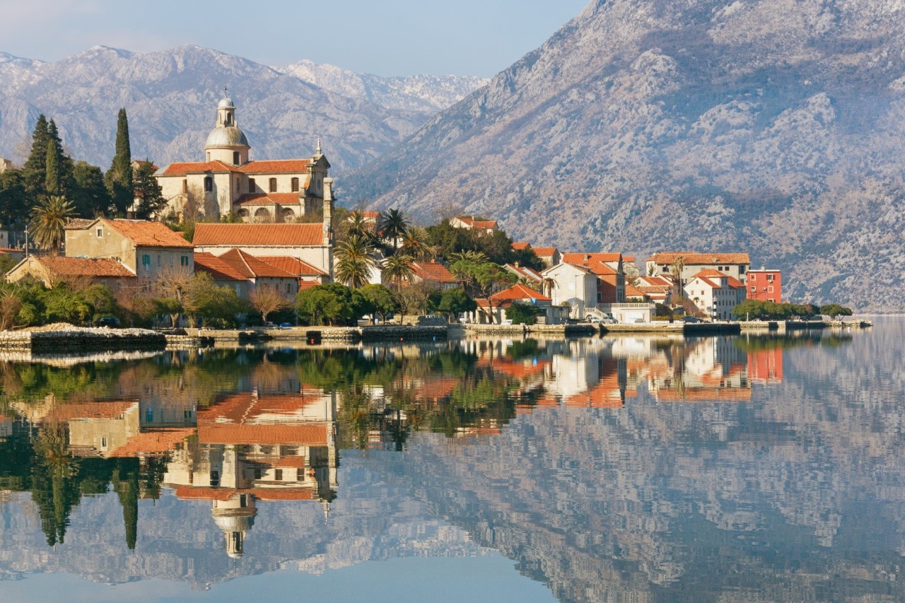 Ort am Wasser. Im Vordergrund spiegeln sich die roten Ziegeldächer, eine imposante Kirche mit Kuppel und die umliegenden Gebäude im stillen Gewässer. Im Hintergrund erheben sich hohe felsige Berge.