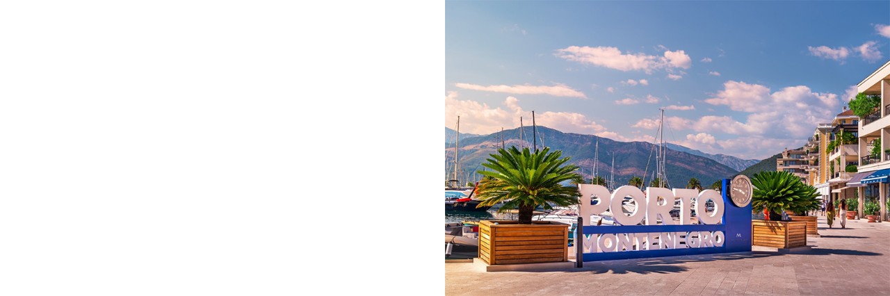 großes Schild mit einer darin integrierten Uhr und der Aufschrift „PORTO MONTENEGRO“. Dahinter erstreckt sich das Wasser mit Segelbooten, eingerahmt von einer Bergkulisse. Rechts verläuft eine Uferpromenade mit Pflanzen und modernen Häusern. 