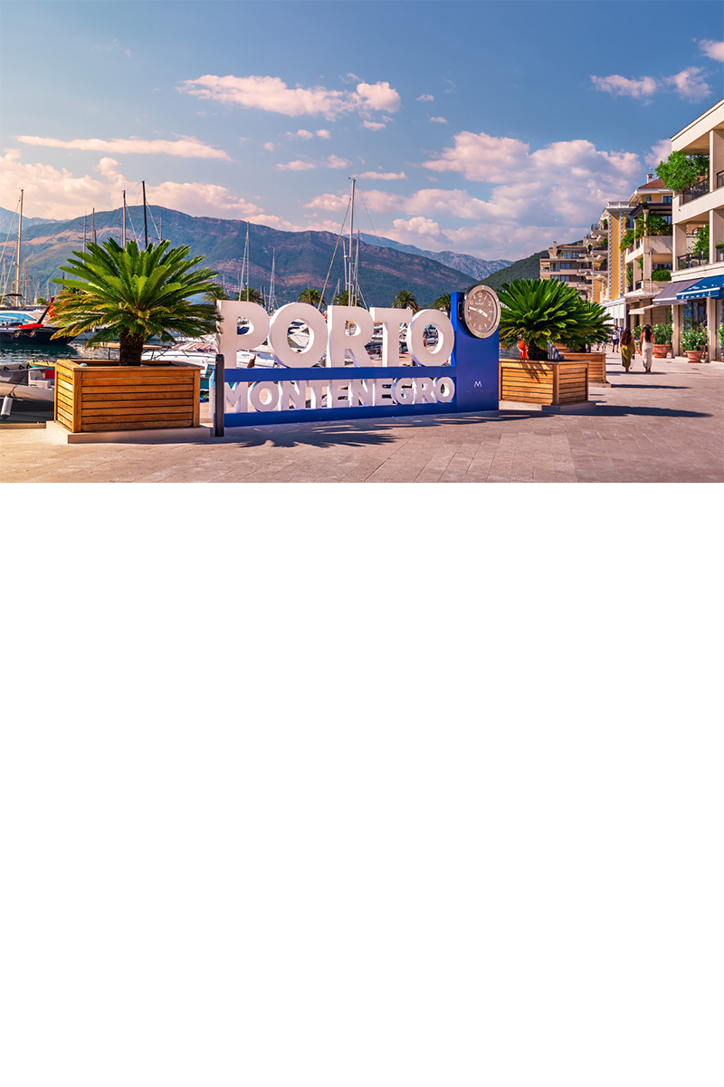 großes Schild mit einer darin integrierten Uhr und der Aufschrift „PORTO MONTENEGRO“. Dahinter erstreckt sich das Wasser mit Segelbooten, eingerahmt von einer Bergkulisse. Rechts verläuft eine Uferpromenade mit Pflanzen und modernen Häusern. 