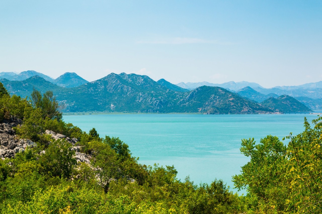 Türkisblauer See, der im Vordergrund durch grün bewachsene Felsen und im Hintergrund von einer hügeligen Landschaft abgegrenzt ist.