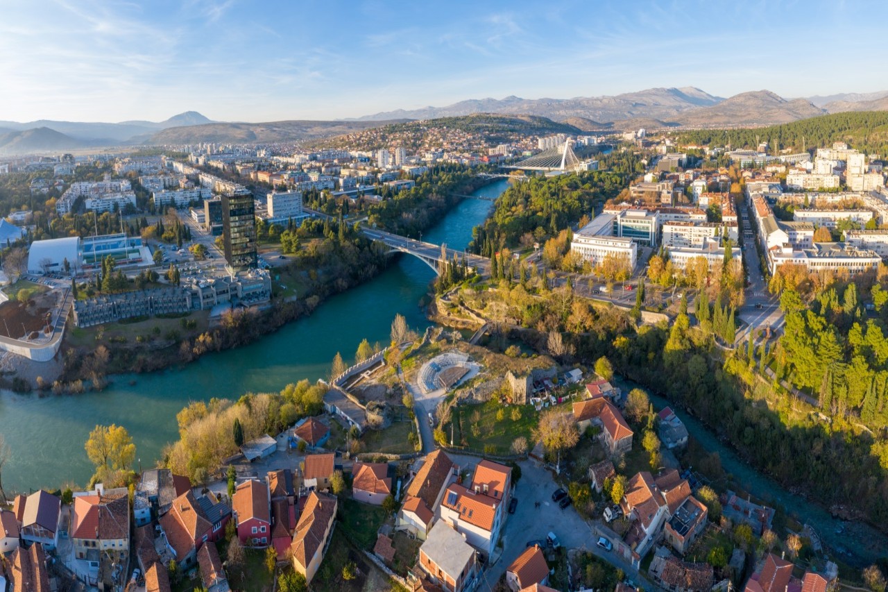 Luftbild einer Stadt, die von einem Fluss geteilt wird, von dem ein weiterer Flussarm abgeht. Über den großen Fluss führen zwei Brücken. Die Flussufer sind dicht bewaldet. Im Vordergrund sind Häuser mit Ziegeldächern sichtbar. Im Hintergrund zeichnet sich eine Hügellandschaft ab.