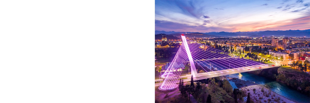Pink angestrahlte Schrägseilbrücke im Abendlicht, die zwei Flussufer einer Stadt miteinander verbindet. An beiden Flussufern leuchten die Häuser und Straßen der Stadt, der Abendhimmel strahlt in vielen Farben.