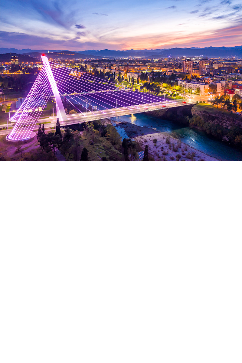 Pink angestrahlte Schrägseilbrücke im Abendlicht, die zwei Flussufer einer Stadt miteinander verbindet. An beiden Flussufern leuchten die Häuser und Straßen der Stadt, der Abendhimmel strahlt in vielen Farben.