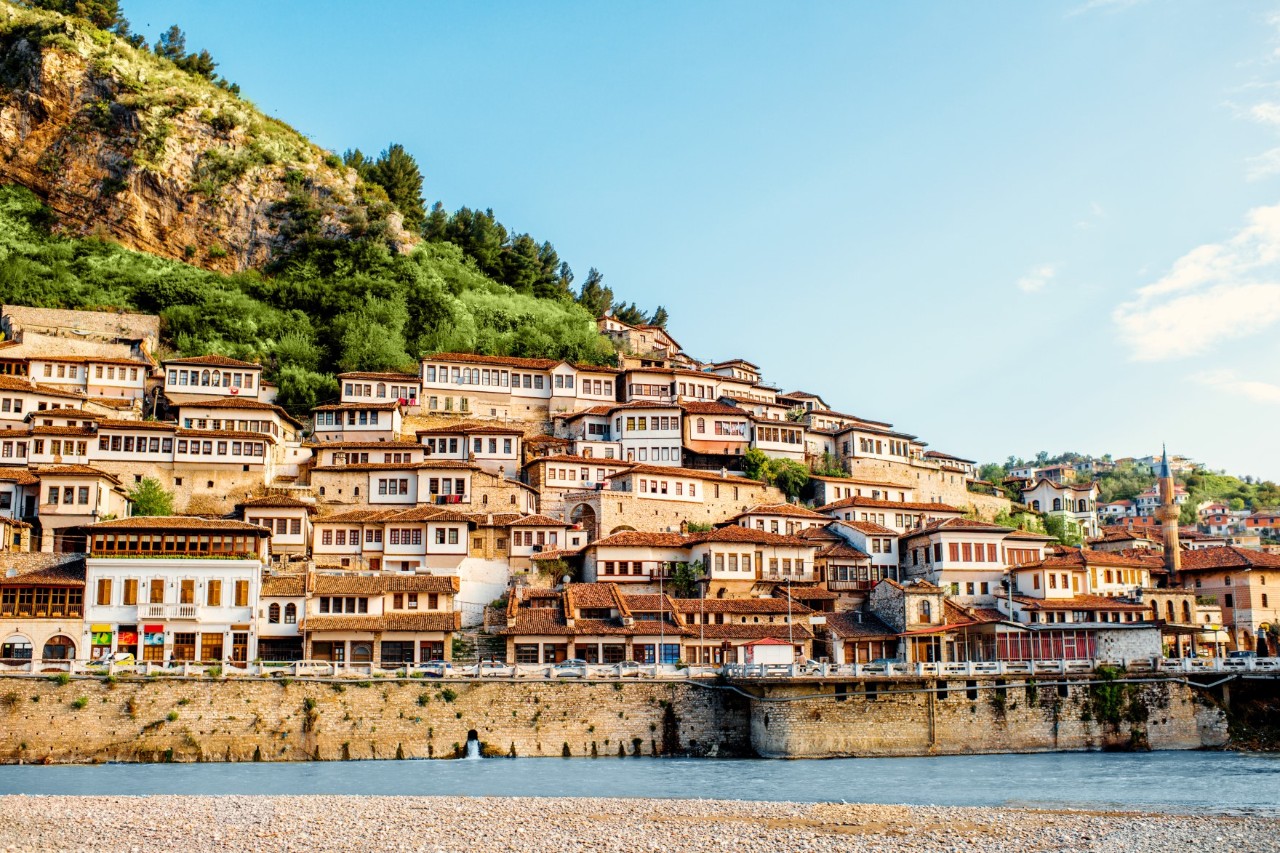 Historische Stadtansicht mit zahlreichen weißen Häusern und roten Dächern, die terrassenförmig am Hang angeordnet sind. Jedes Haus besitzt viele Fenster. Im Vordergrund fließt ein Fluss.