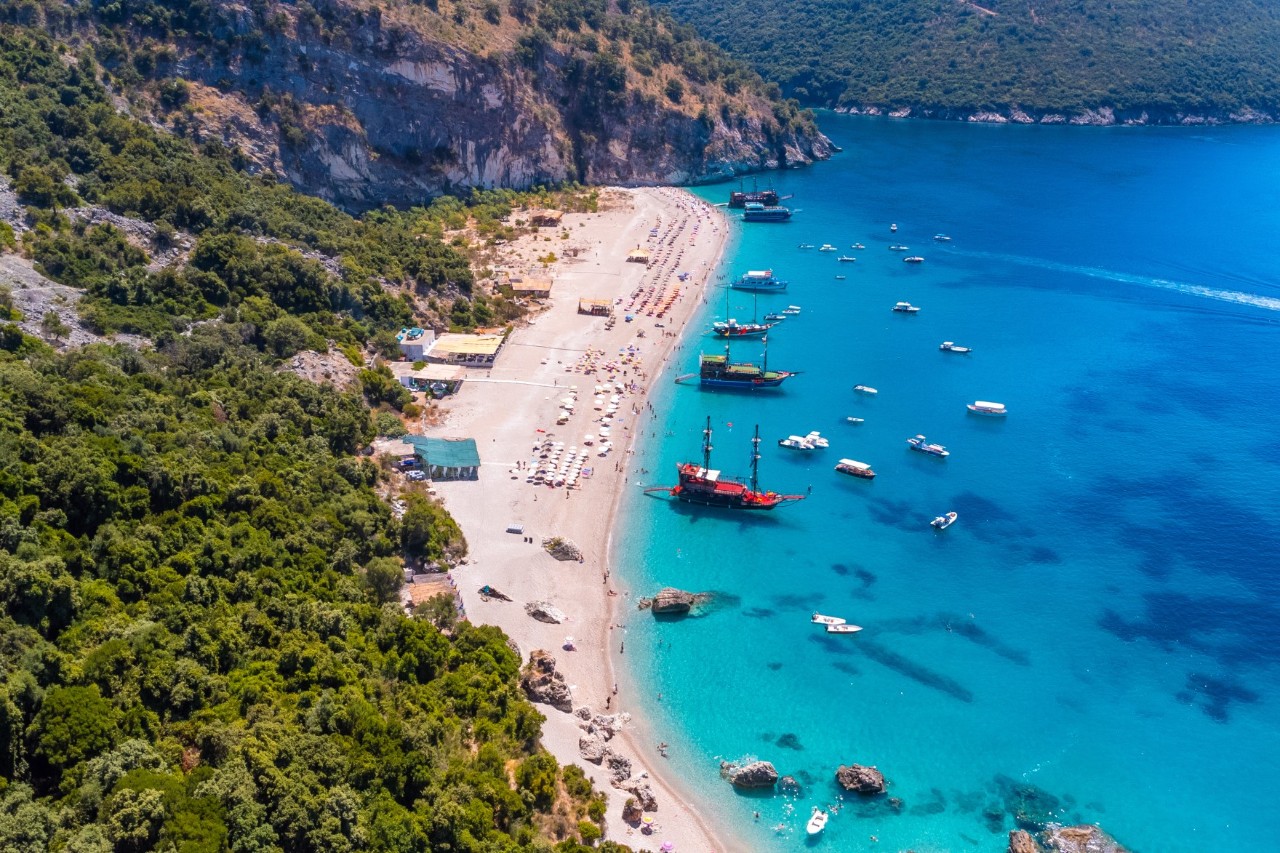 Luftaufnahme eines Sandstrands am blauen, kristallklaren Meer. Am Ufer ankern zahlreiche Boote. Am Strand sind Häuser und Sonnenschirme zu sehen. Die Bucht wird von einer felsigen Küste mit grüner Vegetation eingerahmt.