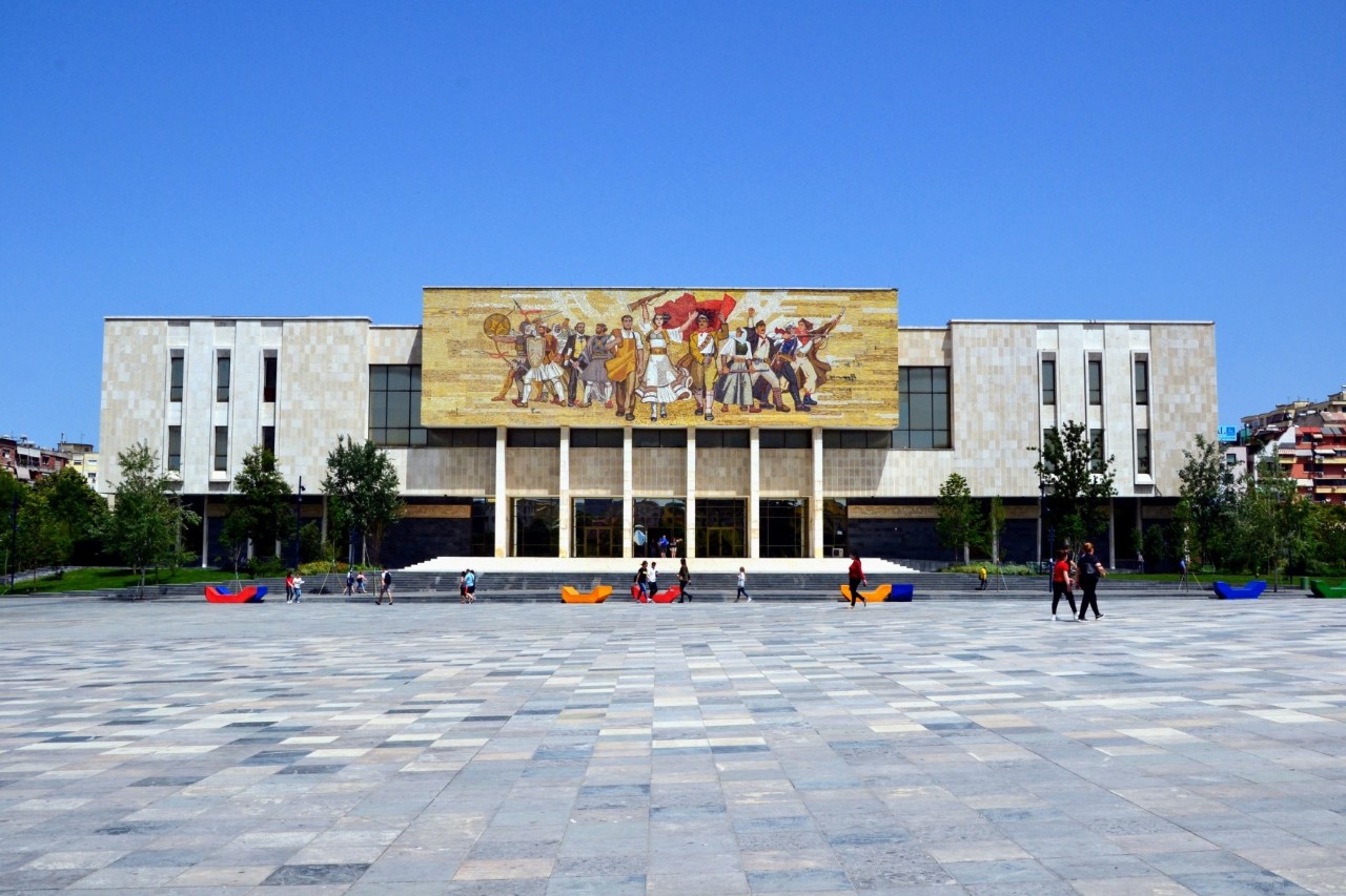 Großes Gebäude mit breiter Fassade. Über dem Eingang ist ein imposantes Mosaik zu sehen, das eine Gruppe von Menschen darstellt. Davor erstreckt sich ein Platz mit wenigen Menschen und bunten Bänken.