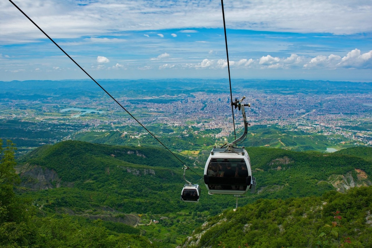 Seilbahn mit zwei Kabinen, die über eine grüne, bergige Landschaft fährt. Im Hintergrund erstreckt sich eine weite Stadtansicht bis zum Horizont.