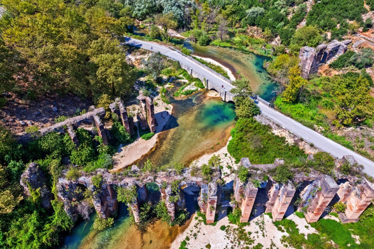 Vogelperspektive auf eine grüne Landschaft mit einer alten, teils verfallenen römischen Aquäduktbrücke, die über einen Fluss führt. Daneben verläuft eine moderne Straße.