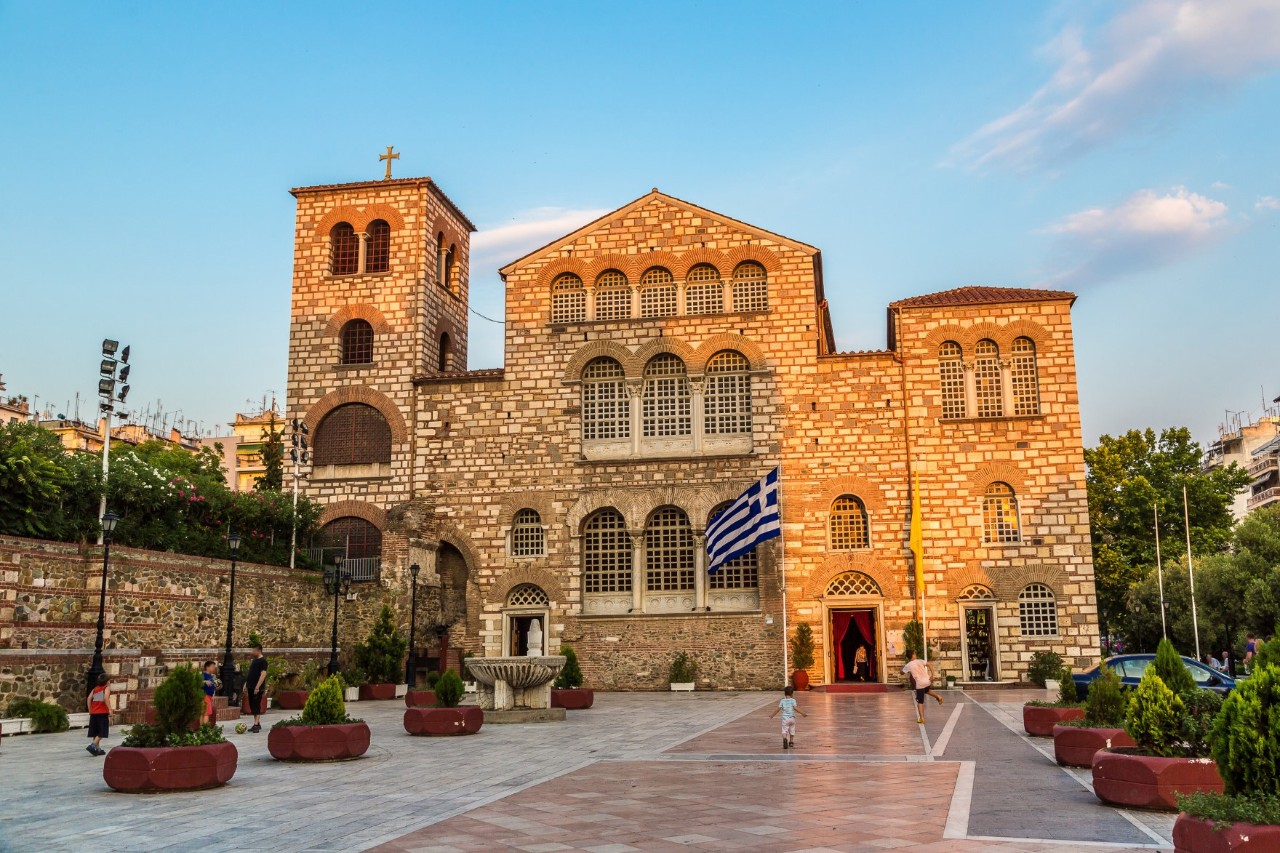 Eine byzantinische Kirche aus Backstein mit großen, verzierten Fenstern und einem Glockenturm zu sehen. Vor der Kirche befindet sich ein großer Platz mit Pflanzen und wenigen Menschen.