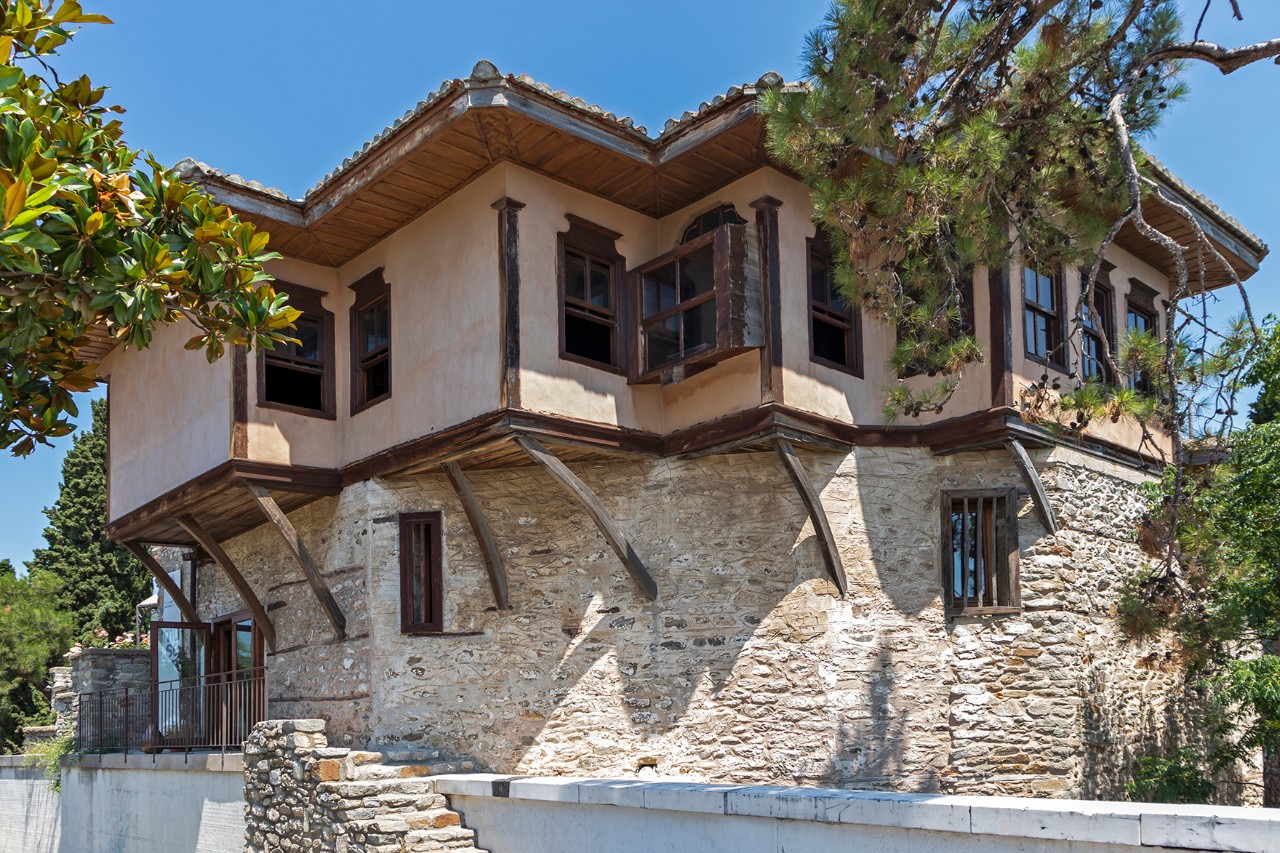 Historisches Wohnhaus mit traditioneller osmanischer Architektur in der Altstadt Panagia. Das zweigeschossige Gebäude besitzt hölzerne Balkone und helle Fassaden und ist umgeben von Bäumen.