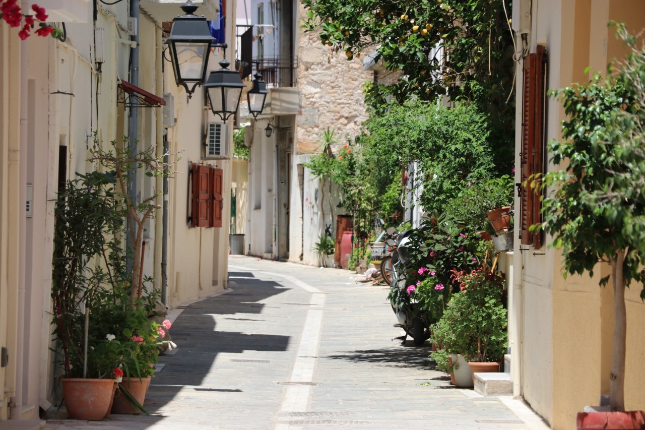 Mediterrane Gasse. Links und rechts im Bild sind sandfarbene Häuserfronten, mit Laternen, braunen Rollläden. Davor stehen viele Blumenkübel.