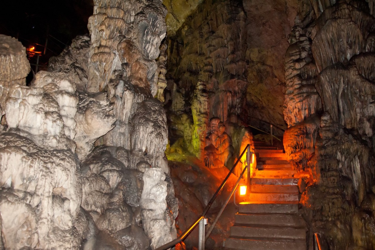 Das Innere einer Höhle mit Tropfsteinformationen. In der Mitte führt eine Treppe hinauf, welche beleuchtet ist. 