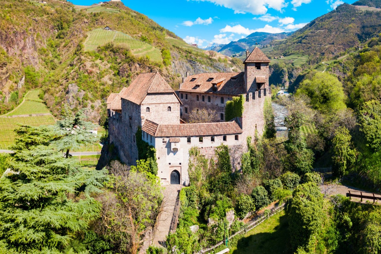 Großer Burgkomplex mit Ziegeldächern, Zinnen zu einer Seite und einem eckigen Turm in einer Berglandschaft. Ein Weg führt zu einem Bogeneingang in die Burganlage. Rund um die Burg ist Wald zu sehen, dahinter erheben sich Berge.