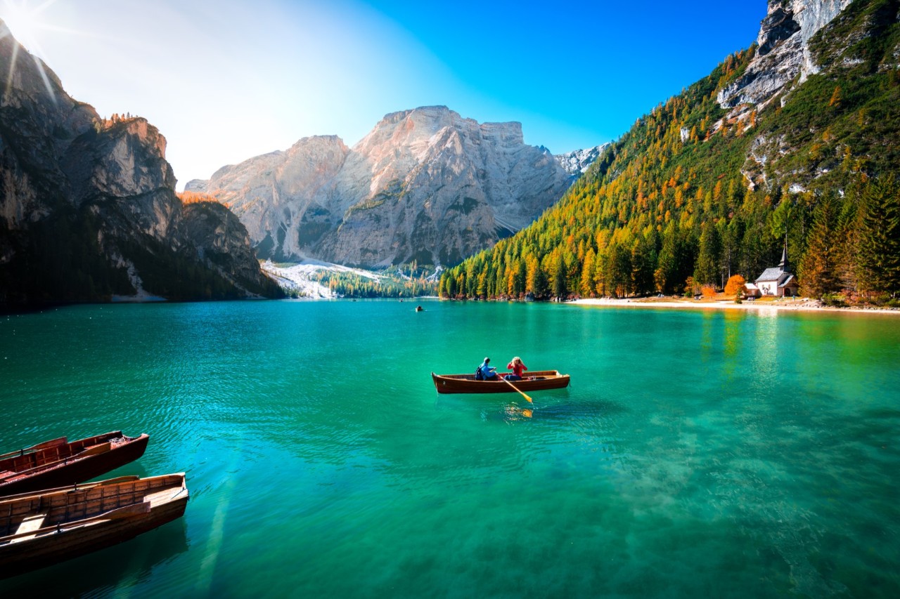 Smaragdgrüner See, zu allen Seiten erheben sich hohe Berge. Auf dem See ist ein Holzruderboot mit zwei Menschen zu sehen, im Bildvordergrund ankern zwei weitere Holzboote in Ufernähe. Am rechten Ufer ist ein kleines Häuschen sichtbar.
