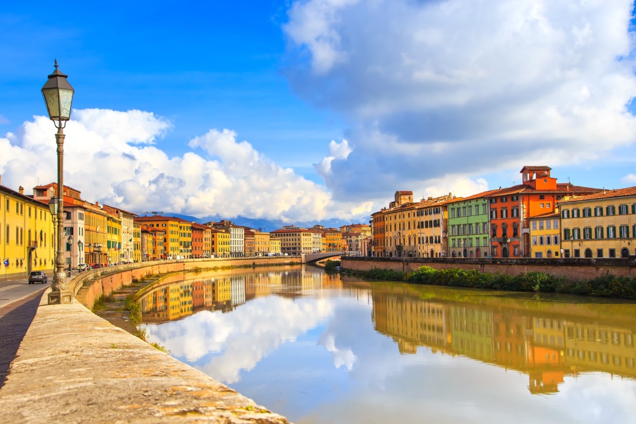 Der Fluss Arno in Pisa, der gesäumt ist von bunten Altbauhäusern, die sich bei Sonnenlicht auf der Wasseroberfläche spiegeln. Auf der linken Bildseite steht eine hübsche alte Straßenlaterne 