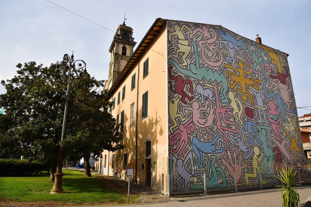 Ansicht der bunten Wandmalerei an der Fassade einer Kirche in Pisa. Links neben der Kirche steht ein Baum 