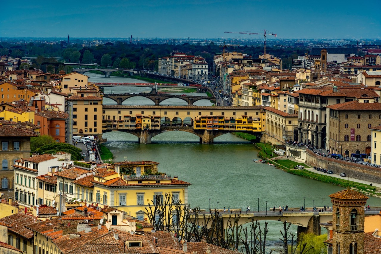 Stadtansicht von oben. Zentral im Bild befindet sich ein breiter Fluss, über den Bogenbrücken führen. Mittig des Flusslaufs ist die Brücke Ponte Vecchio, die mit Häusern bebaut ist 