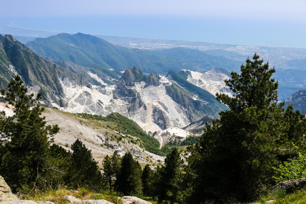 Eine Bergkette und teils abgebaute Steinbrüche. Im Vordergrund sind Nadelbäume zu sehen 
