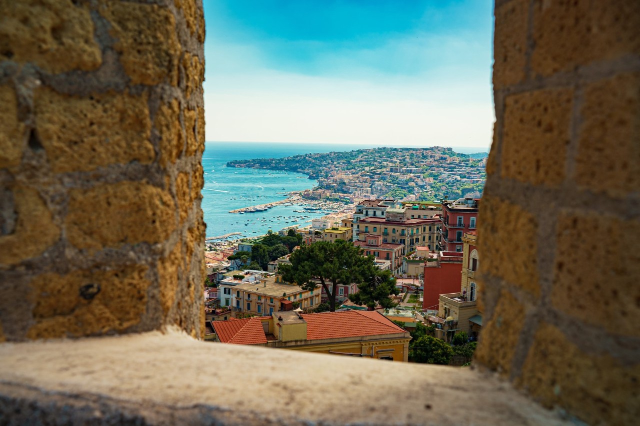 Ein offenes, von alten Mauern eingerahmtes Fenster auf das unten liegende Neapel, die Küste und das Meer.  