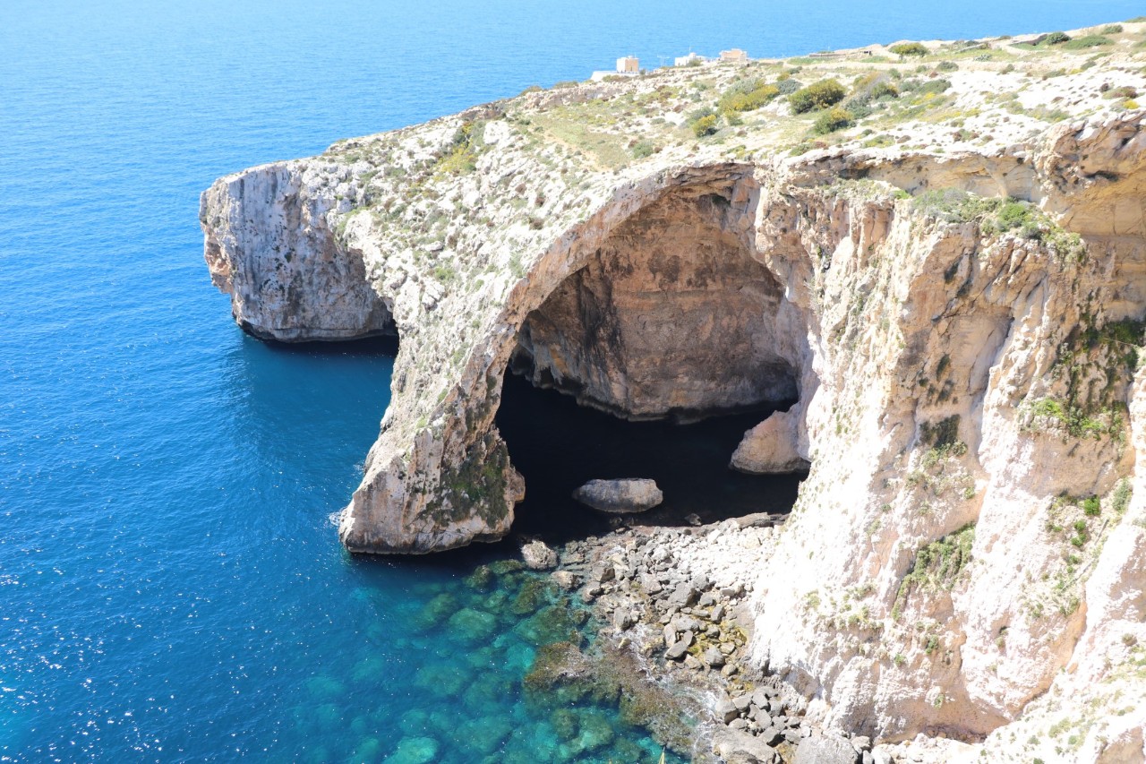 Sandsteinfarbene Felsformationen mit karger Vegetation, die ins Meer ragt. 