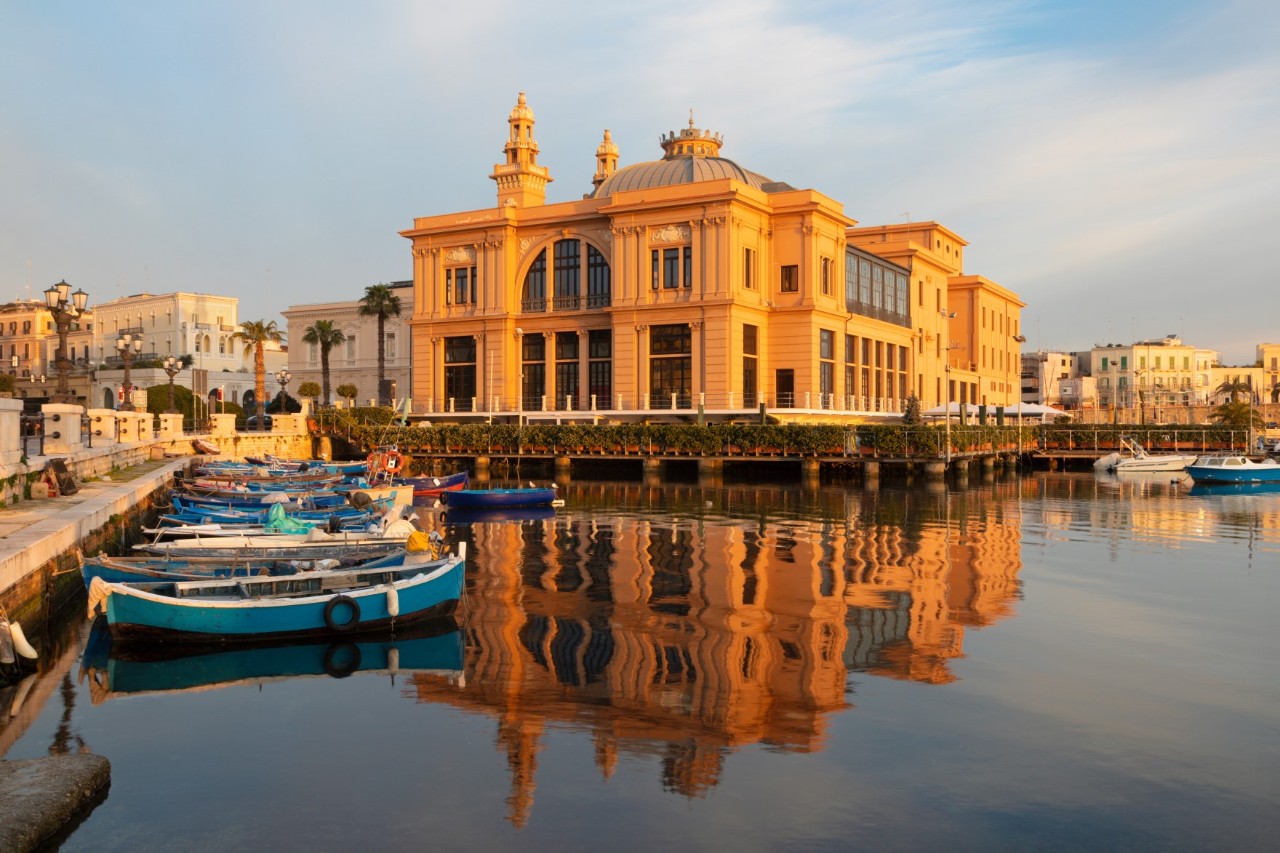 Ein großes im Jugendstil gebautes markantes Gebäude erscheint in der Abendsonne orange. Am Ufer des Gewässers davor liegen Boote.