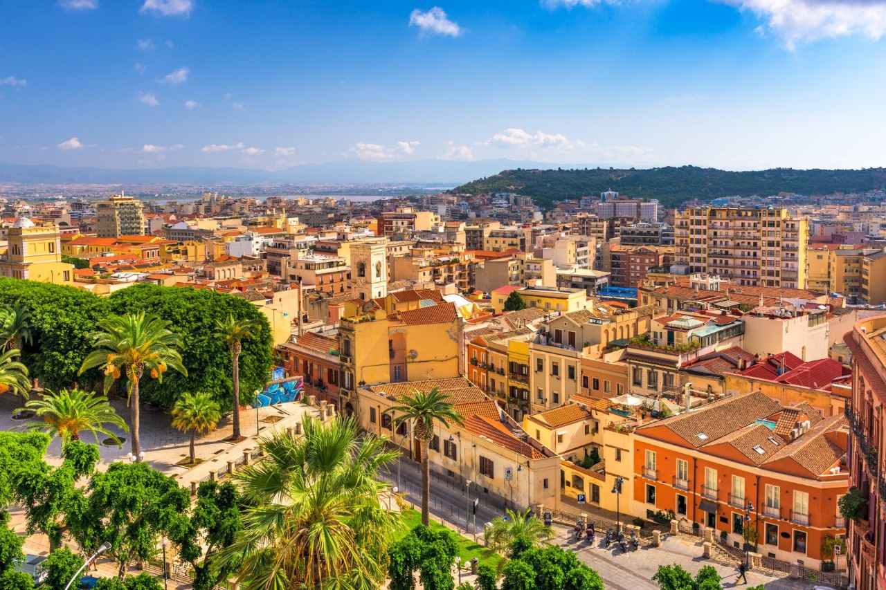 Mediterrane Stadt mit farbenfrohen Gebäuden und dicht aneinandergereihten Dächern bei Sonnenschein. Im Vordergrund erstreckt sich eine grüne Parklandschaft mit Palmen. Im Hintergrund breitet sich die Stadt bis hin zu grünen Hügeln aus.