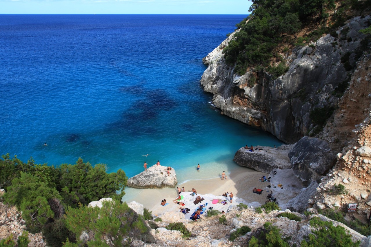 Blick von oben auf eine kleine Bucht mit türkisfarbenem Wasser, umgeben von steilen Felswänden und dichter Vegetation. Auf dem weißen Sandstrand und den umliegenden Felsen sind Menschen zu sehen. 