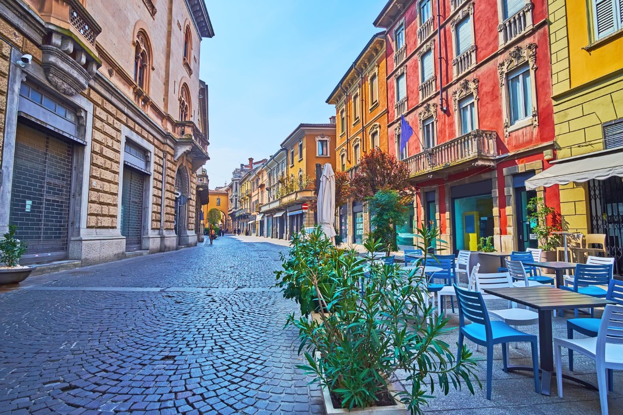 Mediterraner Straßenzug mit Kopfsteinpflaster. Links und rechts davon sind historische Gebäude mit roten, gelben und beigefarbenen Fassaden zu sehen. Rechts vorn stehen bepflanzte Kübel, blaue und weiße Stühle mit Tischen vor einem Lokal.
